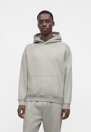 Man wearing a light gray hooded sweatshirt and matching sweatpants standing against a plain white background.