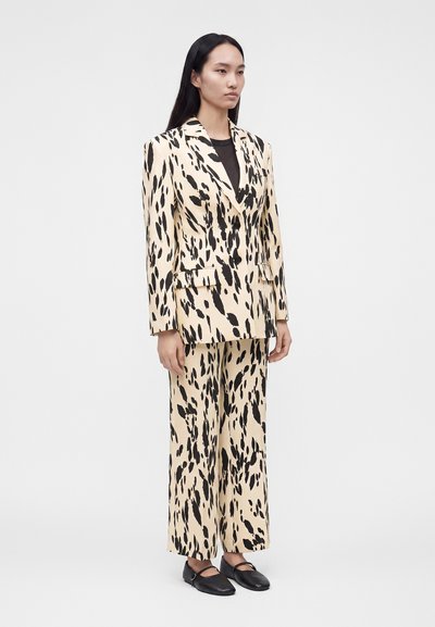 Femme debout portant un blazer beige assorti et un pantalon large avec des taches abstraites noires, associé à des chaussures plates noires.