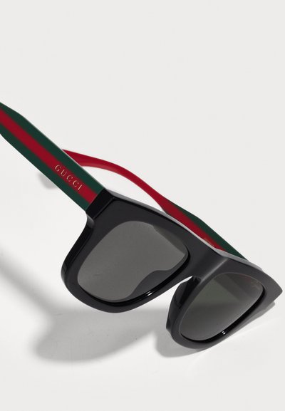 Óculos de sol pretos com lentes escuras, armação quadrada larga e temples listrados em verde-vermelho-verde, apresentando a marca "GUCCI" em dourado.
