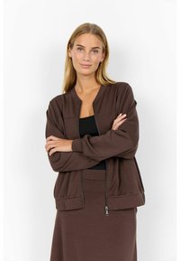 Frau mit langen blonden Haaren, die eine braune Zip-Jacke über einem schwarzen Top trägt, Arme verschränkt, stehend vor einem weißen Hintergrund.
