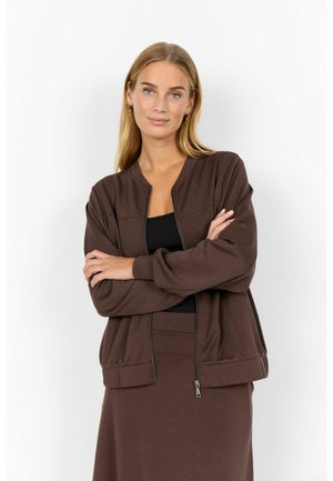 Frau mit langen blonden Haaren, die eine braune Zip-Jacke über einem schwarzen Top trägt, Arme verschränkt, stehend vor einem weißen Hintergrund.