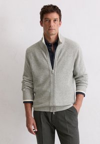 Grau gerippter Reißverschluss-Pullover mit hohem Kragen und kontrastierendem weißen Saum an den Bündchen, kombiniert mit dunkelgrauen Hosen. Strukturiertes Strickmaterial.