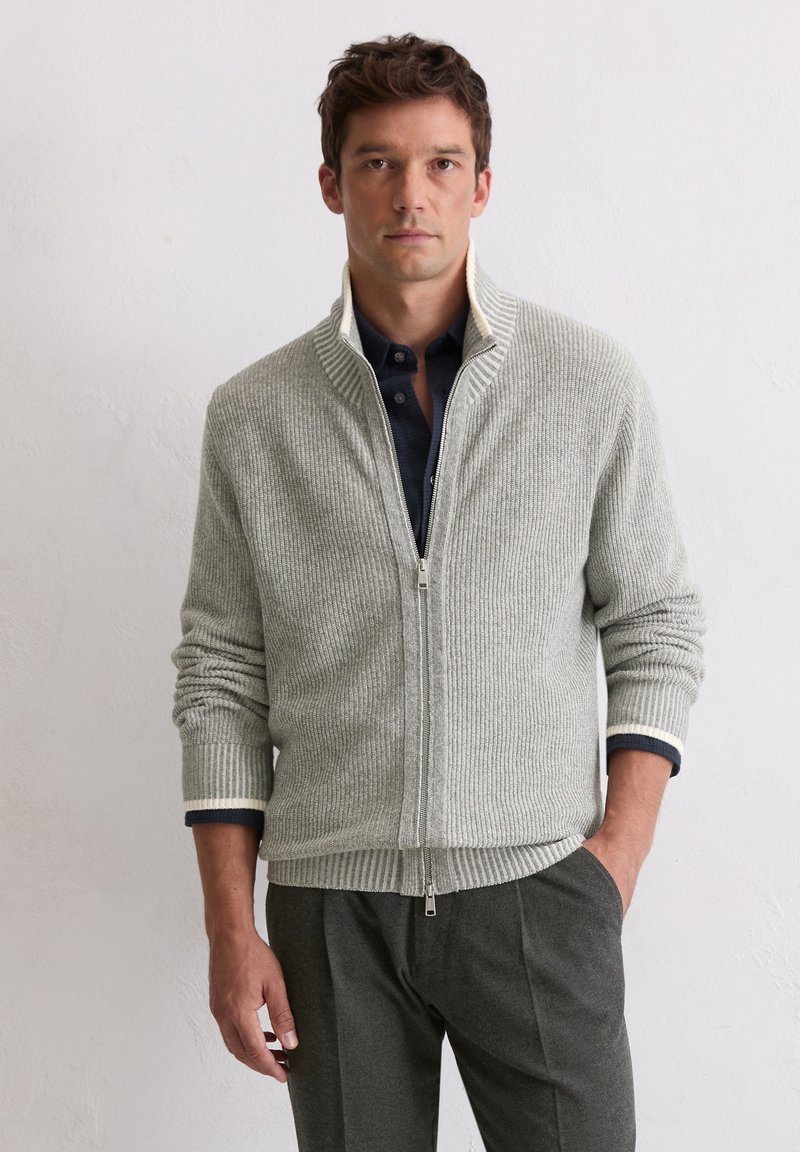 Grau gerippter Reißverschluss-Pullover mit hohem Kragen und kontrastierendem weißen Saum an den Bündchen, kombiniert mit dunkelgrauen Hosen. Strukturiertes Strickmaterial.