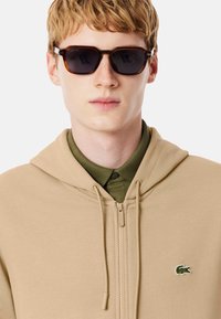 Sudadera beige con cremallera y capucha ajustable con cordón, con logo de cocodrilo verde, llevada sobre una camisa verde con cuello. Gafas de sol oscuras completan el conjunto.