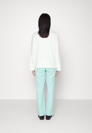 Kobieta stoi twarzą do białej ściany, ubrana w biały długi rękaw, jasnoniebieskie spodnie i czarne buty, z prostymi czarnymi włosami opadającymi luźno.