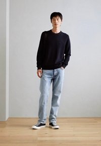 Schwarzer Pullover, hellblaue Jeans, schwarz-weiße Sneakers. Einfaches Design mit glatter Textur; stehende Pose vor einem neutralen Hintergrund.