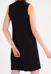 Musta ilmanhiha dressi korkealla kaula-aukolla, sileä kangas, istuva muoto ja hienovarainen takasauma. Ei näkyviä kuvioita tai yksityiskohtia.