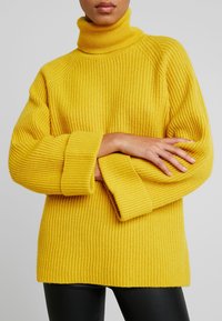 Personne portant un col roulé côtelé jaune vif avec des poignets repliés, bras croisés, associée à un pantalon noir sur un fond gris uni.
