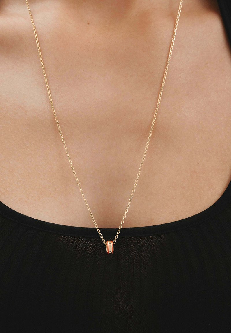 Collar de cadena de oro que presenta un colgante circular con una superficie lisa y pulida, descansando sobre piel desnuda encima de una blusa negra.