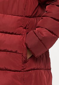 Rdeča napihnjena jakna z kviltanim dizajnom in stransko žepom. Tkanina ima gladko, sijočo teksturo, kar poudarja njen izolacijski videz.