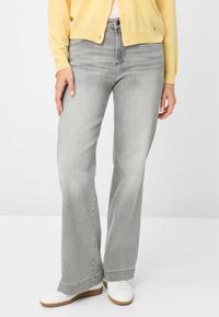 Jeans grigi a zampa con lavaggio chiaro, dotati di chiusura frontale con bottoni e quattro tasche. Abbinati a sneaker bianche e un cardigan giallo.