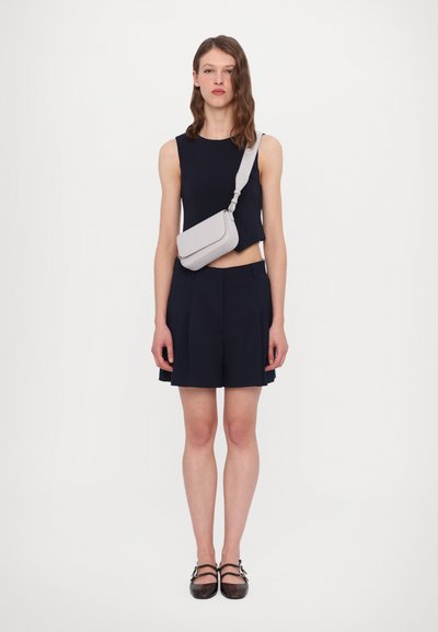 Top senza maniche blu scuro con orlo corto, pantaloni corti sartoriali e borsa a tracolla grigia chiara. La modella indossa scarpe marroni con cinturini.