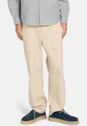 Beige cotton trousers with a loose fit, featuring two large front pockets, contrast stitching, and paired with dark blue suede shoes.