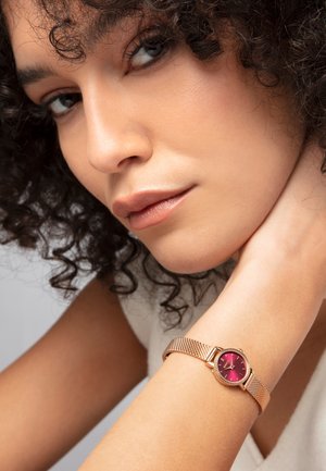 Montre en maille rose doré avec un cadran rond rouge. Présente des index horaires minimalistes et un bracelet texturé. Teint de peau visible sur le poignet.