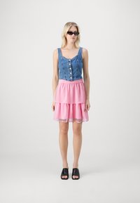 Denim topp med knapper og rosa prikker lagdelt skjørt. Svarte åpne mules. Modellen har på seg solbriller og står foran en hvit bakgrunn.