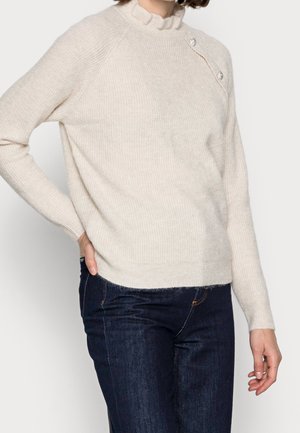 Pull en maille beige clair avec une texture côtelée, un col à volants haut, et trois boutons décoratifs sur l'épaule. Associé à un jean foncé.