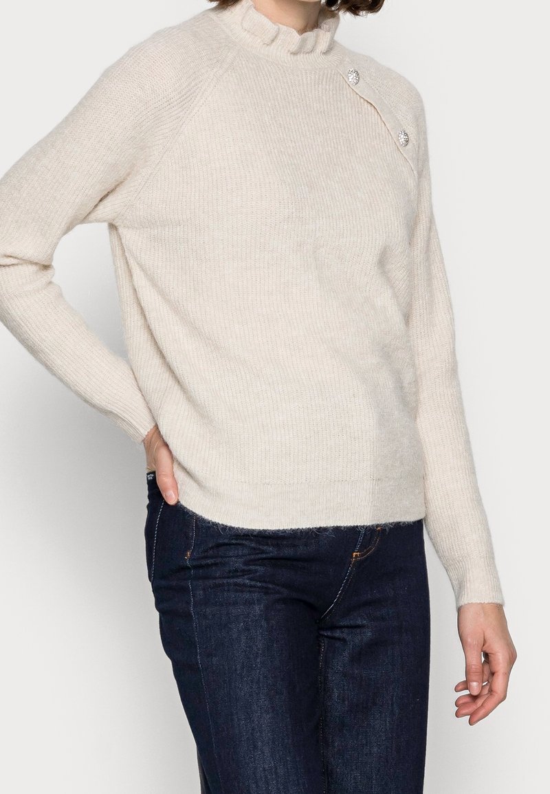 Pull en maille beige clair avec une texture côtelée, un col à volants haut, et trois boutons décoratifs sur l'épaule. Associé à un jean foncé.