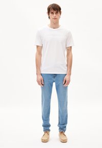T-shirt en coton blanc avec un texte de marque subtil, associé à un jean en denim bleu clair et des chaussures de style mocassin beiges. Fond uni.