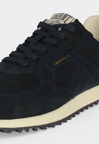 Baskets en daim bleu foncé avec lacets noirs, semelle blanche et logo "GANT" jaune sur le côté, montrant une partie de l'étiquette de la languette.