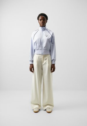 Chaqueta corta azul claro con ribete oscuro y logo bordado, combinada con pantalones anchos blancos y zapatillas beige. Diseño simple y contemporáneo.