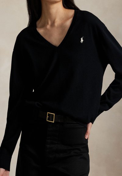 Woman wearing a black V-neck sweater with a white embroidered logo on the chest, tucked into black pants with a belt.