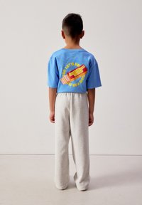 Enfant debout, tourné de dos, portant un t-shirt bleu avec un texte graphique coloré et un pantalon ample gris clair sur un fond uni.