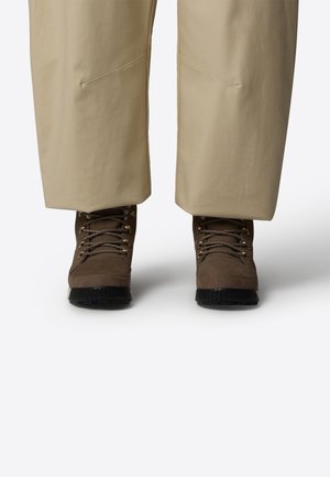 Brune udendørsstøvler med en tekstureret overflade, snørelukning foran, metallic øjer og en robust sort gummisål. Parret med lyse beige bukser.