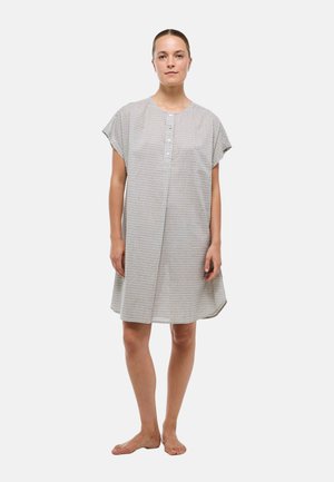 Femme debout pieds nus portant une robe grise à carreaux, ample, à manches courtes et boutonnée, sur un fond uni.