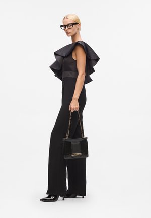 Schwarzer Jumpsuit mit einem rüschierten Satin-Schulterdetail, elegantem Design und taillierter Silhouette, kombiniert mit einer strukturierten schwarzen Handtasche.