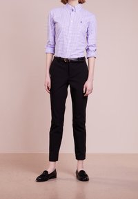 En person iklädd en uppvikt lavendelrutigt skjorta med knappar, svarta slim fit byxor med bälte och svarta loafers stående på ett beige golv.