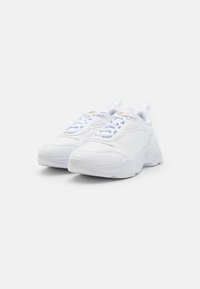 Baskets blanches avec une texture lisse semblable au cuir, forme arrondie, lacets proéminents et semelles rembourrées, présentant un logo discret sur le côté.