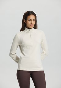 Woman in cream long-sleeve zip-up top and dark brown pants stands with hands behind back against plain background.