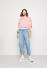 Sweatshirt rosa com fecho éclair e colarinho branco, combinado com jeans de perna reta azul clara e sapatilhas de lona brancas de cano alto.
