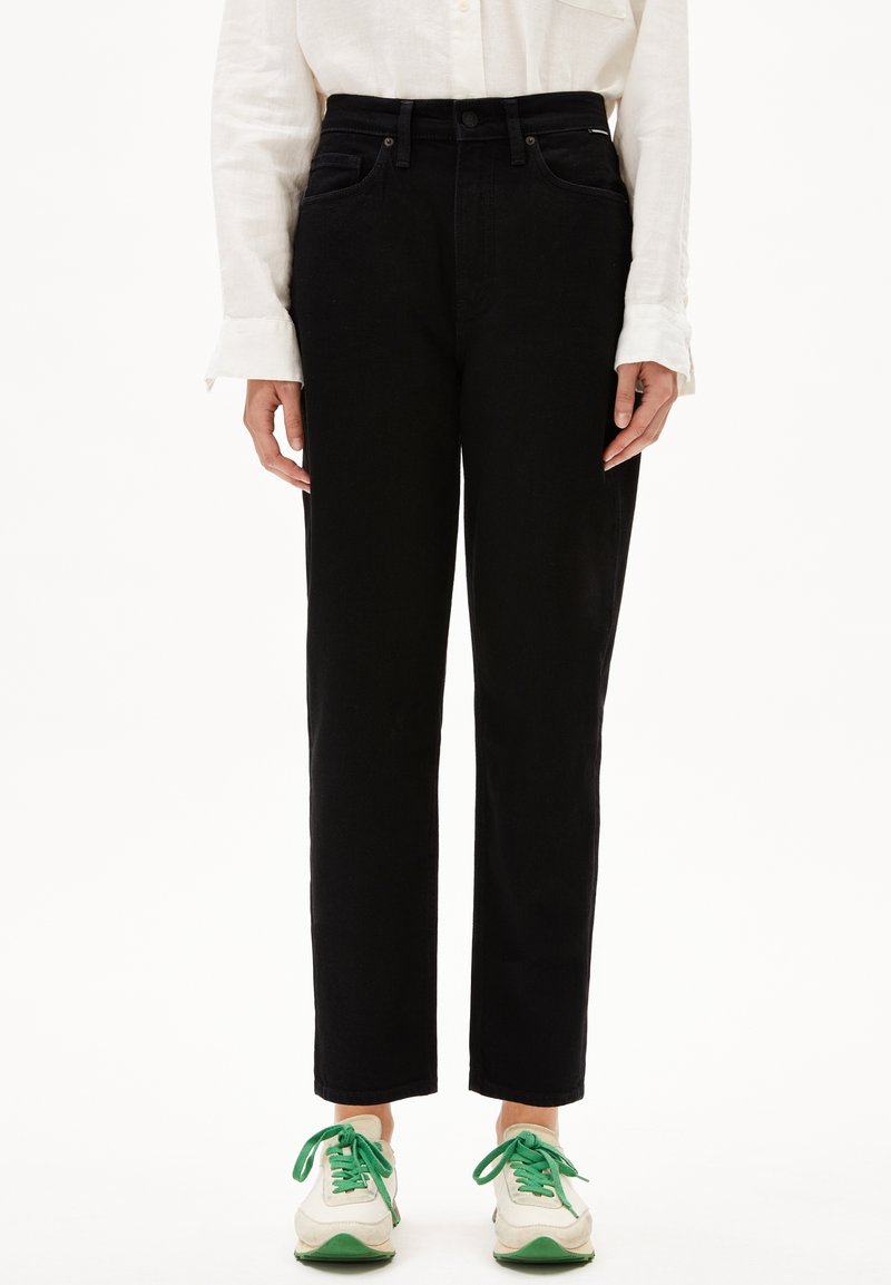 Personne portant un pantalon noir droit, une chemise blanche à manches longues et des baskets blanches avec des lacets et semelles verts, debout devant un fond blanc.