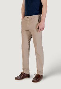 Chino in cotone beige con design a gamba dritta, caratterizzati da due tasche frontali, passanti per cintura e chiusura con bottone. Indossati con scarpe da barca marroni.