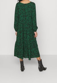 Vestido maxi floral verde con mangas largas, corte relajado y cintura fruncida. Presenta patrones florales en negro. Se combina con botas negras hasta los tobillos.