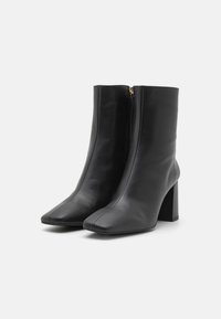 Botas de cuero negro hasta el tobillo con punta cuadrada, cremallera lateral y tacón grueso, con textura suave y un diseño minimalista.