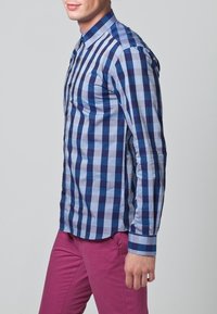 Niebieska i jasnoniebieska koszula w kratę z długimi rękawami, zapinana na guziki z kołnierzykiem, zestawiona z różowymi spodniami.