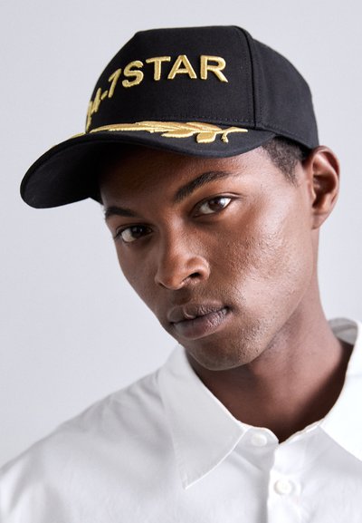 Black baseball cap with gold embroidered text "4-1 STAR" and a leaf design; worn with a white collared shirt.
