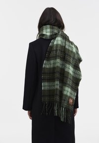 Sciarpa a quadrati verdi realizzata in tessuto testurizzato, con bordi frangiati e una fascetta con logo marrone, esposta su un cappotto scuro.