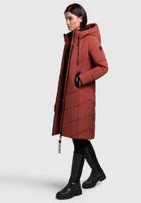 Raudona pūkinė striukė su zigzago raštu, užtrauktuku ir reguliuojama gaubtu. Turi juodus akcentus ir šonines kišenes. Derinama su juodais kelius siekiančiais batais.