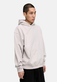 Hellegrauer Hoodie mit geräumiger Fronttasche, Fleece-Textur, überschnittenen Schultern und gerippten Bündchen. Kombiniert mit schwarzen Jogginghosen.