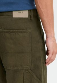 Pantalones cargo verde oliva con superficie texturizada, con una etiqueta de cuero de color claro en la cintura y un detalle de bolsillo lateral.