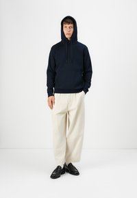 Navy blue hoodie with a front pocket, drawstring hood, and ribbed hem, paired with loose-fitting cream trousers and black loafers.