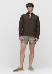 Un homme se tient devant un fond neutre, portant une chemise marron à boutons, un short à motifs et des lunettes de soleil, avec une expression détendue.