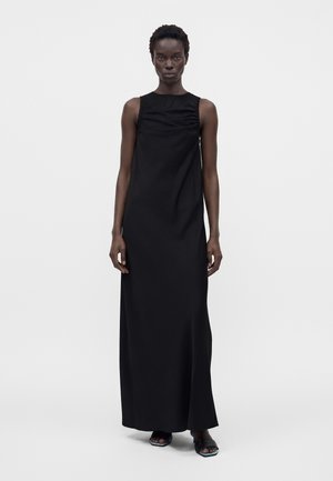 Femme grande aux cheveux courts portant une robe noire sans manches, longue jusqu'au sol, et des sandales noires à bouts ouverts sur un fond clair uni.