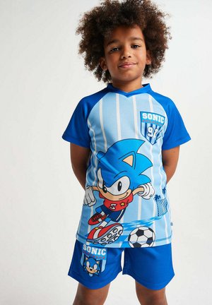 Boy with curly hair wearing blue Sonic-themed soccer jersey and shorts, standing with hands behind back against white background.