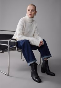 Suéter blanco de cuello alto acanalado, jeans azules cortos con puños enrollados y botines negros de tacón con cremallera lateral, sentada en una silla de metal.