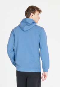 Blå hoodie, mjuk bomullsblandning, känguruficka, justerbar huva. Ärmar och nederkant är ribbade. Baksidan visar sömnadsdetaljer och rena linjer.
