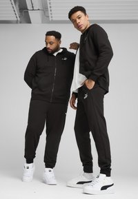 Felpe sportive nere con cappuccio e pantaloni jogger, caratterizzati da loghi bianchi Puma, abbinati a sneakers bianche. Tessuto liscio e design dalla vestibilità rilassata.